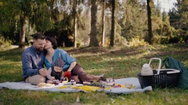 Yavrusu olan genç bir aile, yeşil ağaçlar ve piknik sepetiyle çevrili sakin bir orman ortamında piknik yaparken neşe içinde eğleniyor. Genç Aile Güneşli Orman Parkı 'nda Piknik Yapıyor