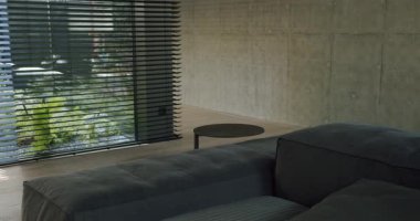 Gösterişli, modern bir oturma odasında koyu gri bir kanepe, yuvarlak bir sehpa ve beton duvarlar bulunur. Geniş pencereler dışarıdaki yeşil yeşillikleri gösteriyor, iç konforu açık hava tazeliğiyle harmanlıyor..