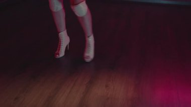 Pembe ve mor ışıklı loş bir odada dans eden yüksek topuklu kadın bacakları. Yeteneğini ve dans tutkusunu sergileyerek özgüven ve zarafetle hareket ediyor. Kadın Şehvetli Dans