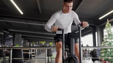 Kendini adamış genç bir adam iyi donanımlı bir fitness merkezinde hava bisikleti üzerinde çabalıyor, azim ve zindelik sergiliyor. Modern Spor Salonu 'nda Odaklanmış Sporcu Hava Bisikleti Çalışıyor