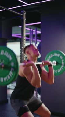 Kararlı bir erkek sporcu modern bir spor salonunda halterle çömelme hareketi yapar. Gücünü ve forma bağlılığını gösterir. Spor salonlarının parlak mor ışıkları egzersizine dinamik bir enerji katıyor..