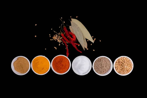 Six Round Bowls In One Line Filled With Spices, Dried Red Chilies With Seeds And Cinnamon Leaves. Isolated On Black Background. Spices Like, Mustard Seeds, Coriander Seeds, Cumin Seeds, Red Chili Powder, Turmeric Powder And Salt. Copy Space