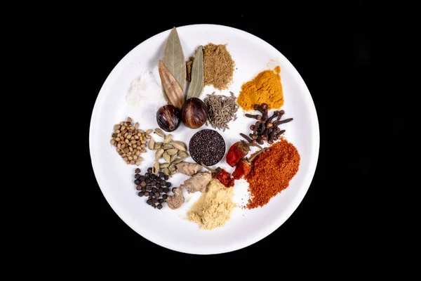 Top View Of White Dish Filed With Many Spices Like Spices Like, Mustard Seeds, Coriander Seeds, Cumin Seeds, Red Chili Powder, Turmeric Powder, Salt, Black Pepper, Clove, Cardamom Or Elaichi, Hing, Raw Turmeric And Nutmeg. Isolated On Black Backgroun