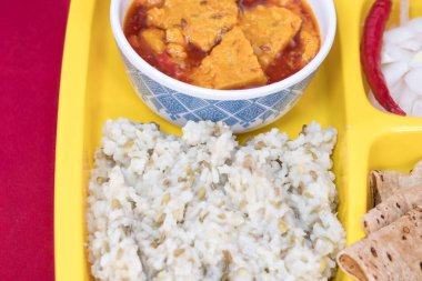 Very Close-Up view of Gujarati Kathiyawadi Spicy Dhokali Sabji, khichdi, Roti, Onion Pieces, Red Chilli, on yellow square dish prominently, set against a red background clipart