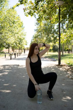 Spor kıyafetli genç bir bayan sporcunun vücudu bir şişe suyla parktaki spor sahasında oturuyor ve başka tarafa bakıyor.