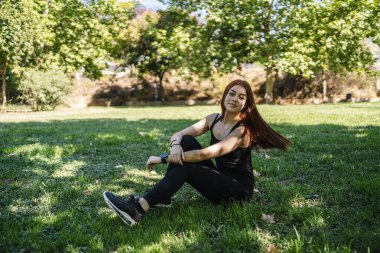 Parkta çimlerin üzerinde oturmuş kameraya bakan sıradan giyinmiş genç bir kadın.