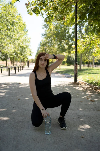 Spor kıyafetli genç bir bayan sporcu parkta asfalt yolda oturuyor ve kameraya bakıyor.