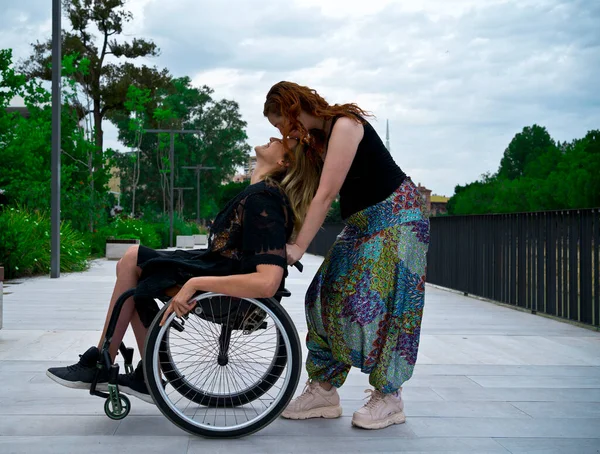 Full body side view of young lesbian woman standing behind back of girlfriend using wheelchair and kissing on forehead during stroll in park