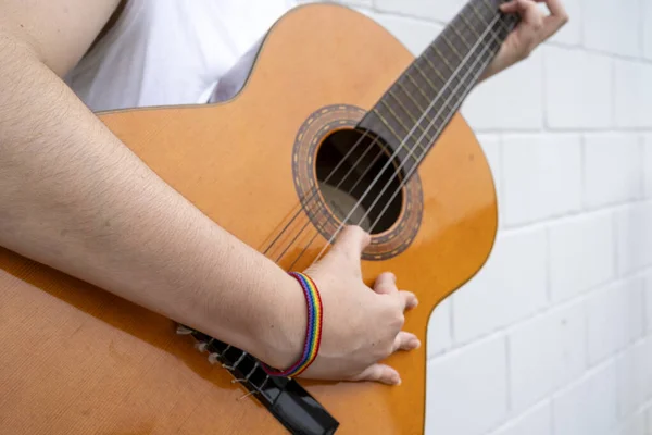 Crop unrecognizable female guitar player with rainbow LGBT bracelet playing music on acoustic guitar and performing while standing against white tiled wall
