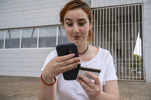 Delighted woman in casual clothes standing on street and checking data from credit card on smartphone screen