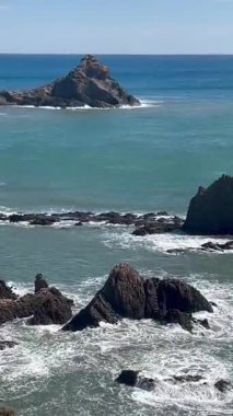 Güçlü turkuaz dalgalar karanlık kayalık kıyı şeridine doğru yükseliyor. Geniş mavi gökyüzünün altında el değmemiş okyanus ufkuyla birlikte uzak bir adayı ortaya çıkarıyor.