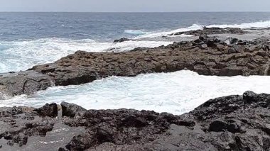 Beyaz köpüklü okyanus dalgaları tenerife, kanarya adaları, İspanya sahillerindeki karanlık bir volkanik kaya oluşumuna ritmik olarak çarpıyor ve büyüleyici bir doğal güç ve güzellik gösterisi yaratıyor.