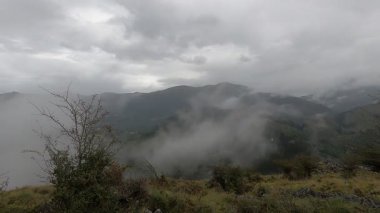 Bulutlar zarif bir şekilde bir dağ sırasının üzerinden akıyor, zaman zaman aşağıdaki canlı yeşil vadiyi gözler önüne seriyor, nefes kesici bir doğa gösterisi yaratıyor.