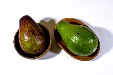 İki yeşil avokado mini ahşap bir tabakta ve bir tanesi de küçük tahta bir kasede sade beyaz arka planda sıralanır.