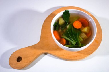 içinde havuç, patates ve hardal sebze çorbası bulunan porselen bir kase, ahşap bir kesme tahtası ve sade beyaz bir sırt kepçesi üzerinde servis edilir.