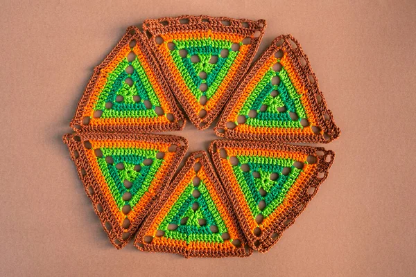 Round figure made of crochet triangles in green and orange tones on beige background. Concept of creative crocheting.