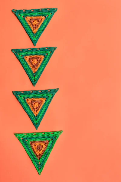 Top view on green crochet triangles with orange parts, placed vertically in a row on vivid orange background. Geometric crochet motifs.