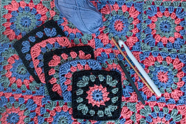 Top view of granny squares with black boardes, crochet hooks of different sizes and skein of blue yarn on texture of multicolored crochet afghan.
