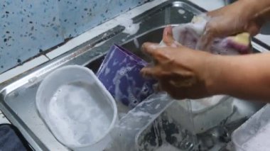 Bulaşıkları sabunla yıkayan bir adam, akan suyun altında tabak ve kap kacak yıkıyor. Sabun köpükleri ve su akışı temiz ve hijyenik bir sahne yaratır. Temizlik ve ev işleri için mükemmel bir tema. 4K