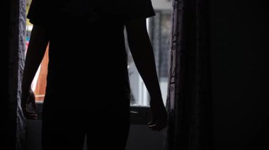 Sabah yatak odasının perdelerini açan bir adamın silüeti. Yumuşak ışığın odaya girdiği huzurlu bir an. Sabah rutini, sakin atmosfer, günün yeni başlangıcı.