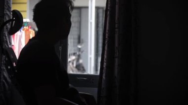 Yatak odasının penceresinin yanındaki sandalyede oturan bir adamın silueti sessizce dışarıya bakıyor ve hayatı yansıtıyor. Huzurlu ve duygusal bir an, yalnızlık ve tefekkür teması için mükemmel. görüntüler 