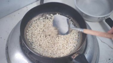 Siyah bir tavada yarı pişmiş yulaf karıştıran bir elin yakın plan görüntüsü. Yemek yaparken yulaflar kalınlaştıkça buhar yavaşça yükselir. Siyah tava, yulafların yumuşak dokusuyla çelişiyor.