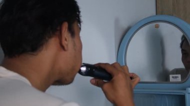 Eskimiş beyaz gömlekli Asyalı bir adam elektrikli tıraş makinesi kullanarak aynanın önünde tıraş oluyor. Kamera, onun odaklanmış duruşunu ve gelişigüzel ayarlarını göstererek sahneyi arkadan yakalıyor. Gerçek bir hayat.