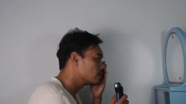 Asyalı bir adam bıyığını jiletle yavaşça tıraş ediyor. Arka plan beyaz ve izole, kamera açısı yandan, yakın çekim bir tımar anı yakalıyor. video görüntüleri 4K. 