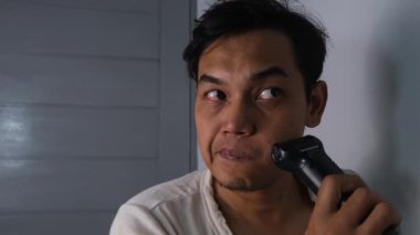 Beyaz gömlekli bir adam elektrikli jilet kullanarak bıyığını tıraş ediyor. Cihazı dikkatle ve dikkatle üst dudağında gezdiriyor. Olay yeri temiz, günlük bakım rutinini vurguluyor. 4K