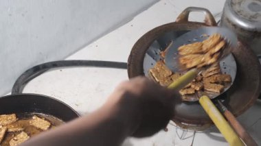 Tempeh 'in yol kenarındaki basit bir yiyecek standında kızarması. Sıcak yağ tavada cızırdıyor. Tempeh 'in altın dilimleri mükemmelliğe ulaşırken. Mütevazı ortam, gelenekselliğin sıcaklığını ve orijinalliğini yansıtıyor. 4K