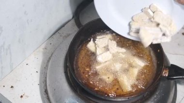 Dilimlenmiş çiğ tempeh sıcak yağ dolu bir tavaya aktarılıyor. Pişirme işlemi, parçalar cızırdayan yağa nazikçe yerleştirilirken başlar. Bu sahne başlangıcı yakalar. 4K