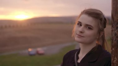 Kahverengi gözlü ve saçlarıyla gülümseyen kız romantik bir yaz günbatımı arka planına sahip bir sütuna yaslanıyor..