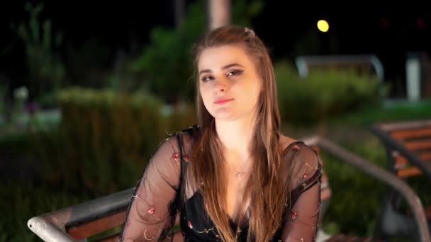 漂亮的年轻姑娘 晚上化着妆坐在灯火辉煌的公园里的长椅上 浪漫的调情情绪 — 图库视频影像