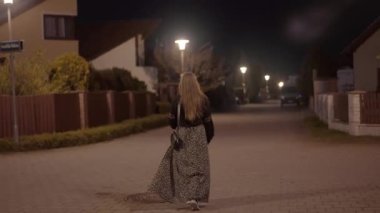 Uzun etekli, dalgalı saçlı bir kız bir gece şehrinde sokak lambalarıyla aydınlatılmış bir sokakta yürüyor.