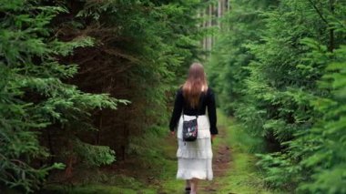 Sırtı dönük bir kadının, yeşil çalılar ve ağaçlar arasında gündüz vakti beyaz etek ve siyah bluz giyerek dar bir yolda yürümesi. Yoğun orman.