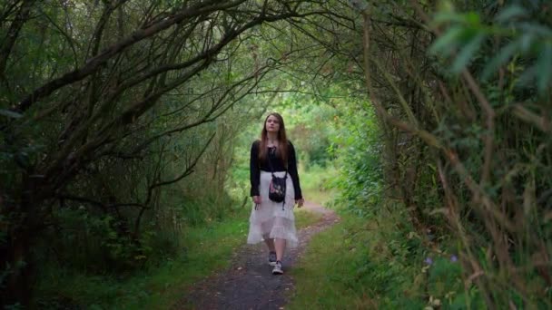 長い髪と白いスカートを持つ美しい若い女性は 木や茂みの緑のトンネルでロマンチックな道を歩いています 晴れた夏の日に静的なスローモーションショット — ストック動画