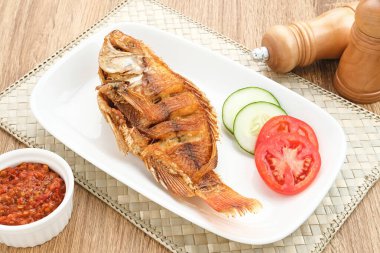 Tilapia ya da Ikan Nila Goreng kızartması. Beyaz tabakta sambal (acılı sos), salatalık ve domates dilimi ile servis edilir.. 