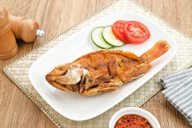 Tilapia ya da Ikan Nila Goreng kızartması. Beyaz tabakta sambal (acılı sos), salatalık ve domates dilimi ile servis edilir.. 