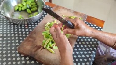 Bir kadının eli yeşil sarmaşık kabağını uzun dilimlerle kesiyor. Mutfaktaki masanın üstündeki tahta tepside bir bıçakla.
