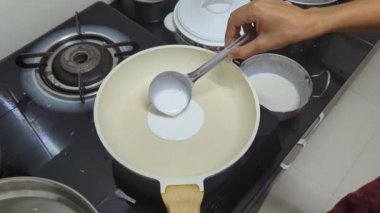Bir kadının eli, beyaz hamuru sıcak bir tavaya doldurur ve kepçeyle mutfakta gaz sobasının altına yuvarlak, lezzetli bir beyaz domates sosu yapmak için yayar.