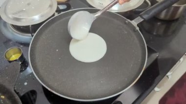 Mutfakta, bir kadın kahvaltıda siyah bir tavaya beyaz hamuru dökerek dosa hazırlar ve kepçeyle sabah mükemmel yuvarlak bir şekle sokar.