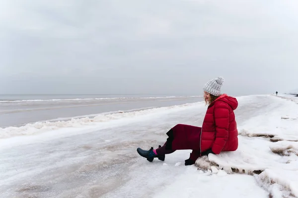 Stock image A cheerful woman in a red jacket and a warm dress sits on the shore of the winter sea.