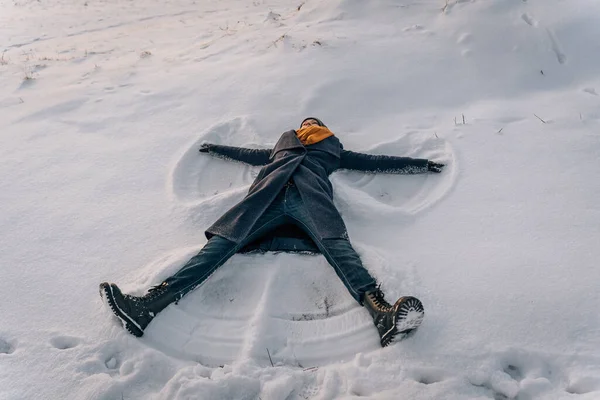 A young woman makes a snow angel lying in the snow with her arms and legs spread to the sides.