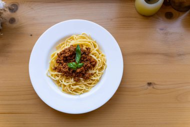 Beyaz bir tabakta spagetti bolonez, ahşap bir masada fesleğen yaprağı ile süslenmiş, yukarıdan görünüyor. Klasik bir İtalyan mutfağı..