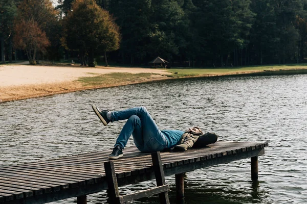 A man lies with his leg behind his leg on a wooden pier on a lake in autumn.