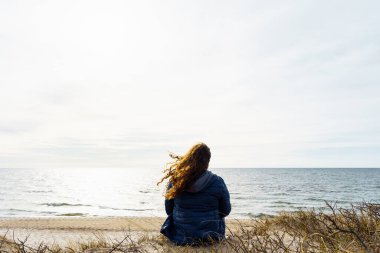 Kıvırcık saçlı, ince ceketli bir kadın akşamleyin deniz kıyısında oturuyor, saçları rüzgarda dalgalanıyor..