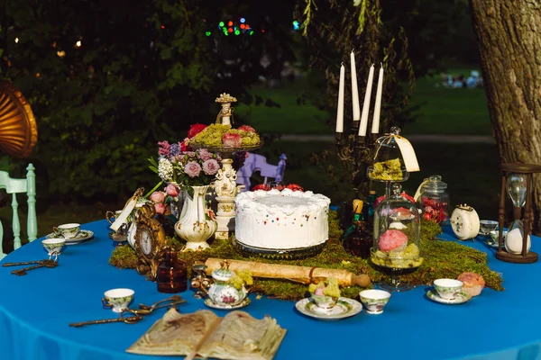 The dining table is decorated in the style of Alice in Wonderland. An open old book, a cake, tea bowls, candles in a candlestick, an hourglass on a blue tablecloth late in the evening.