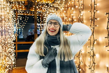 Gözlüklü mutlu genç bir kadın. Kış kıyafetleri içinde neşeli ışıklar altında gülümsüyor..