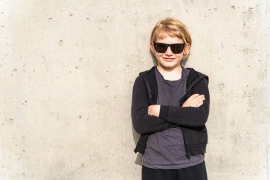 Güneş gözlüklü havalı çocuk beton bir duvara karşı güven içinde poz veriyor..