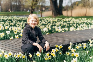 Tahta bir güvertede oturan çocuk çiçek açan nergislerle çevrili, bahar gününün tadını çıkarıyor..